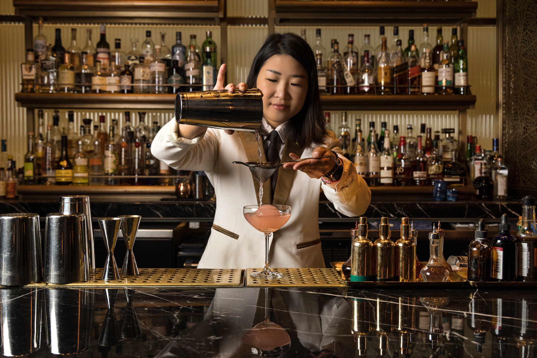 A bartender pouring a pink cocktail into a champagne coupe.