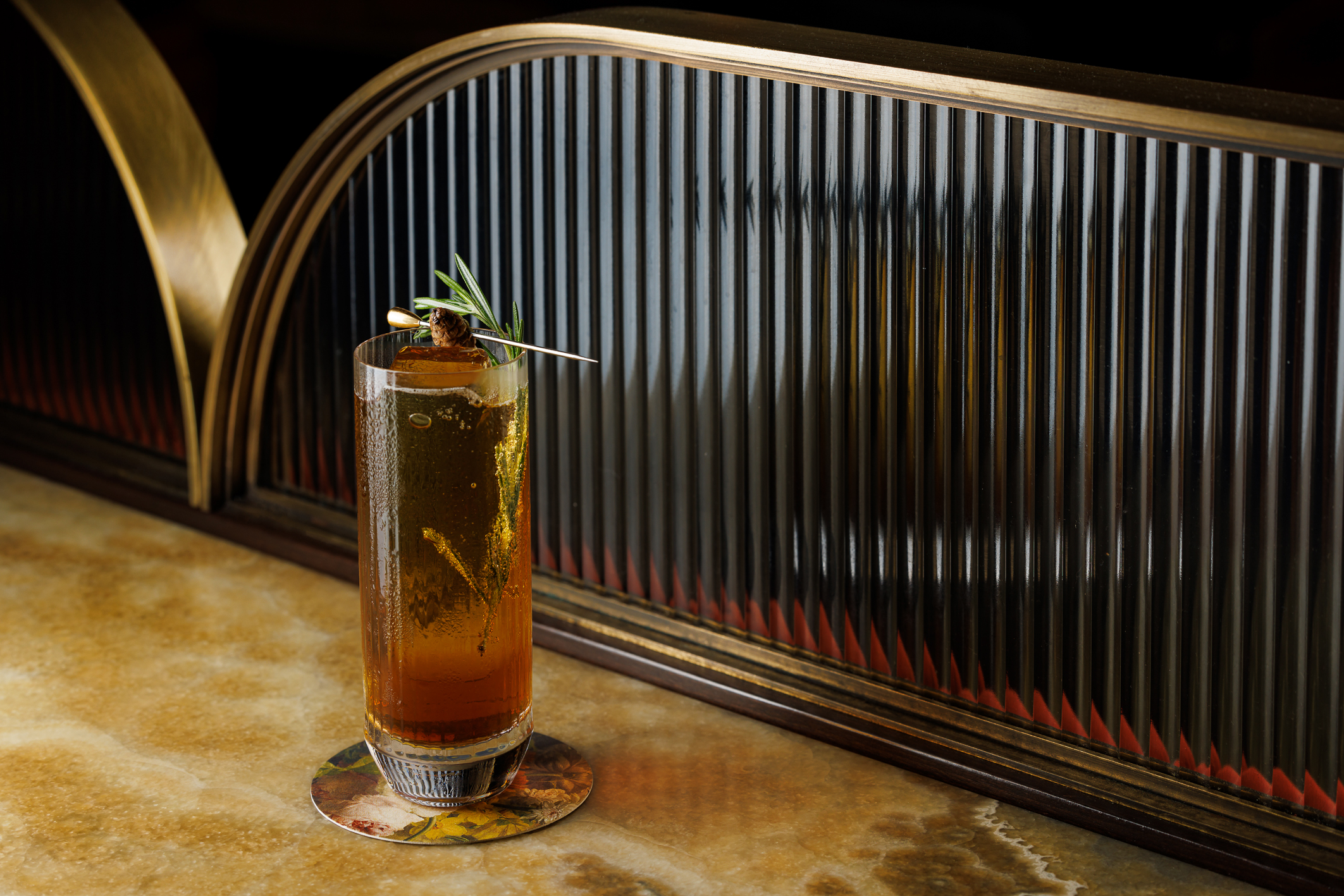 A tall glass with a dark brown cocktail and rosemary garnish.