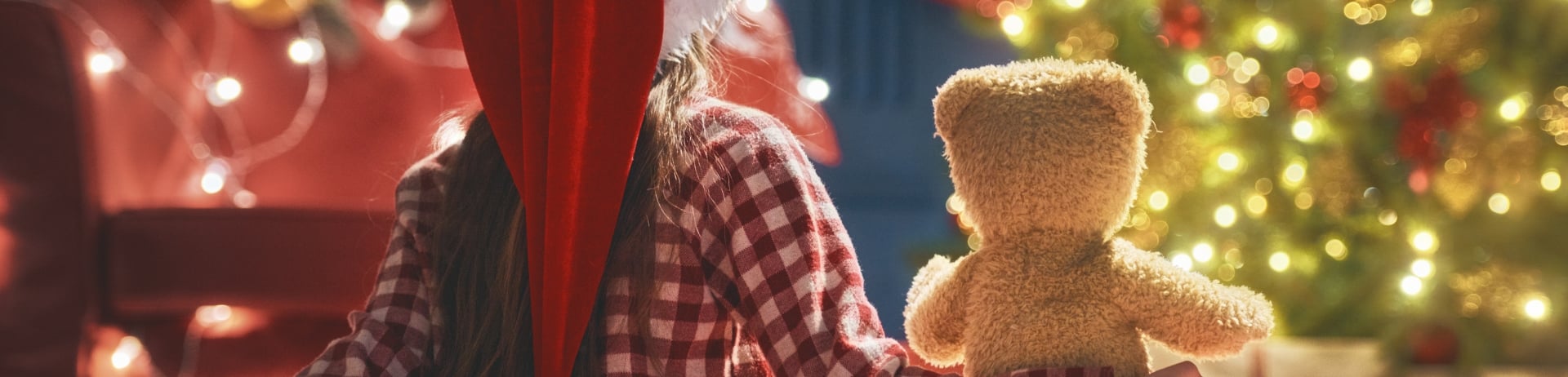 A child sitting in front of a lit Christmas tree holding a teddy bear.