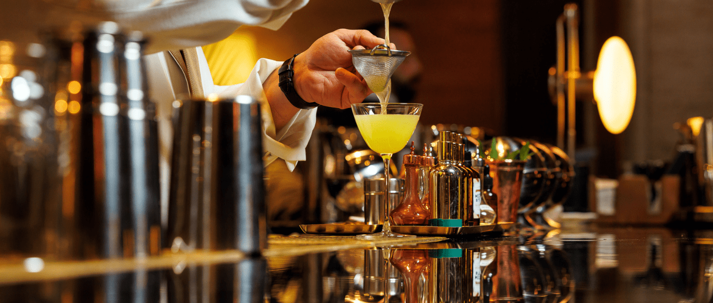 A bartending straining a yellow cocktail through a sieve on the bar top.